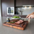 A person slices a medium-rare steak on the BBQ Boards® Best BBQ Board® 16 x 12 Cutting Board in a modern kitchen, with a jar of seasoning, fresh rosemary, and a bowl of salad on the counter.