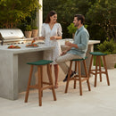 A woman in a white dress serves food at an outdoor kitchen, smiling at a man seated on the Kanyon Living Poly Lumber Saddle Stool Bar Height in Dark Green and Cedar Wood. Surrounded by plants and garden lights, they enjoy a casual meal together.