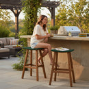 A woman sits on a Kanyon Living Poly Lumber Saddle Stool Bar Height in Dark Green and Cedar Wood at an outdoor stone countertop, holding a drink and smiling, with a grill, plates, and greenery nearby on the sunny patio.