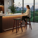 A woman sits on a Kanyon Living Poly Lumber Saddle Stool Bar Height in Dark Green and Cherry Wood at a modern kitchen island, enjoying wine. Bottles and lemons are on the counter. Sunset city views and pendant lights create a warm atmosphere.