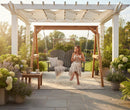 A woman in a light dress relaxes on the Kanyon Living Poly Lumber 4′ Adirondack Swing Deluxe Driftwood Grey under a white pergola, enjoying her mug amid lush plants and sunlight on her peaceful garden patio.