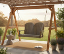 The Kanyon Living Poly Lumber 4′ Adirondack Swing Deluxe Driftwood Grey by Kanyon Living hangs from a pergola on the patio, surrounded by potted plants and an open book on a rug, with a scenic meadow in the background.