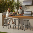 A woman sits on a Kanyon Living Poly Lumber Saddle Stool Bar Height Deluxe in Driftwood Grey, chatting with a man by an outdoor kitchen counter with a grill and drinks. Three of these modern Kanyon Living stools are lined up under the sunny sky.
