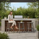 A woman sits on a Kanyon Living Poly Lumber Saddle Stool Bar Height Deluxe in Driftwood Grey by an outdoor kitchen with a grill, surrounded by greenery. Three matching Kanyon Living stools add to the modern, relaxed vibe.