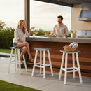 A woman sits on the Kanyon Living Poly Lumber Saddle Stool Bar Height Deluxe in Driftwood Grey and White, chatting with a man behind an outdoor kitchen counter with drinks, fruit, and towels amid coastal seating and lush greenery.