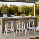 A modern outdoor kitchen with coastal seating features three Kanyon Living Poly Lumber Saddle Stool Bar Height Deluxe Driftwood Grey and White stools at a gray island, plants, a recipe tablet, and a pool with greenery. A person’s arm is partly visible on the left.