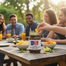 A group of friends relax outdoors with snacks and drinks. In the foreground, a hand dips a chip into a DDR BBQ Supply Five Amigos Fire Roasted Pepper Salsa (Hot), 16 oz jar.