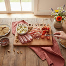 A Words With Boards Seriously Gluten Free Personalized Cutting Board (Small, 9.5"x12.5") by Words with Boards, LLC holds salami, prosciutto, cherry tomatoes, mozzarella balls, walnuts, and jam; a hand arranges tomatoes with tongs beside crackers and flowers.