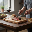 Someone slices a medium-rare steak on a Words With Boards Gluten Free Personalized Cutting Board (6 1/4" x 9 1/2") by Words with Boards, LLC, using a fork and knife. Nearby are a glass of red wine, salad, and bread on the rustic table.