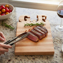A person uses tongs to hold sliced steak on a Words With Boards Gluten Free Personalized Cutting Board (6 1/4" x 9 1/2", Words with Boards, LLC), surrounded by rosemary, cherry tomatoes, red wine, and coarse salt.
