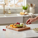 A hand slices blue cheese on the Words With Boards Gluten Free Personalized Cutting Board (6 1/4" x 9 1/2") by Words with Boards, LLC, surrounded by cheddar cubes, grapes, figs, and kiwi in a celiac-safe kitchen with marble counters.