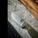 The Spaceman Knives Halimede Fixed Blade Knife, featuring a green Micarta handle and stonewashed D2 steel blade, rests on textured stone near wood. A black sheath with metal rivets from Spaceman Knives is partly visible beside it.