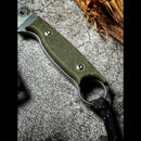 Close-up of the Spaceman Knives Halimede Fixed Blade Knife’s textured green Micarta handle with finger ring, two metal rivets, and a black lanyard, shown resting on a rough gray surface with brown wood in the upper right corner.