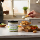 Terrapin Ridge Farms Hatch Chile Cream Cheese Dip sits on a kitchen counter by toasted bread and fresh veggies. A hand dips bread into this irresistible dip, bringing bold Southwest flavor to your snack spread.