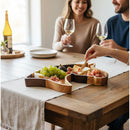 The Wooden Horse Head Serving Tray With Custom Engraving Options by Two Acre Woodworks showcases grapes, figs, cheese, and cured meats on a rustic table as two people smile with wine glasses while a hand reaches for food.