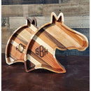 Two Acre Woodworks’ Wooden Horse Head Serving Trays, crafted in varied wood tones, showcase custom monogram engravings at the center. These personalized trays are pictured against a rustic wood-paneled backdrop.