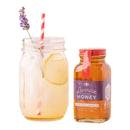 A mason jar glass with iced lemon drink, garnished with a lavender sprig and a red striped straw, sits beside a jar of Honey Truck Co Lavender Infused Honey.