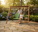 A couple enjoys a peaceful moment on a stone patio in warm sunlight; the woman sits on a lime green and black Kanyon Living Poly Lumber 4′ Adirondack Swing while the man stands beside her, surrounded by blooming flowers and lush greenery.
