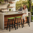 A woman in light clothing smiles at a modern patio counter with a drink, while three Kanyon Living Poly Lumber Saddle Stool Bar Height Lime Green and Black stools line the wooden and concrete surface amid lush greenery.