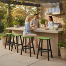 Three people gather around a patio island, smiling and chatting. The space showcases Kanyon Living Poly Lumber Saddle Stool Bar Height in lime green and black, potted plants, a grill, and a counter filled with drinks and food.