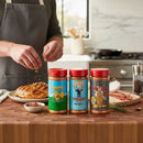 A person sprinkles Meat Church Southern Sweet Smoke Pack rubs on cooked meat in a kitchen. Three colorful Meat Church spice jars with red lids are lined up on a wooden counter beside herbs, bacon, and a small bowl of spices.
