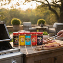 Ten vibrant Meat Church Seasoning bottles from the Meat Church 10 Pack sit on an outdoor table by a grill, while someone uses tongs to place seasoned steaks onto the grates in a sunny backyard.