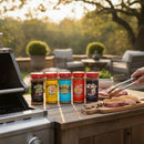 Ten vibrant Meat Church Seasoning jars from the 10 Pack line up on an outdoor kitchen counter near a grill, as someone uses tongs to season raw steaks. Patio furniture and greenery appear in the sunny background.