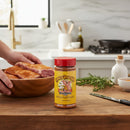 A jar from the Meat Church Seasoning 10 Pack by Meat Church sits on a kitchen counter, while hands season raw meat in a wooden bowl with herbs and additional BBQ rubs visible nearby.