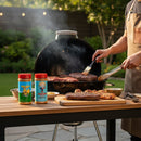 A person grills meat on a round charcoal grill in the backyard, using tongs and a basting brush. The Meat Church Southern Sweet Smoke Pack by Meat Church and cooked meat sit on a wooden table; green trees and string lights are in the background.