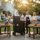 Friends gather at sunset for a backyard barbecue, smiling and chatting around a Myron Mixon Smokers Myron Mixon Gravity Fed Smoker G20, with food and drinks on nearby tables amid greenery and glowing string lights.