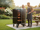 A man and woman smile in their backyard by a Myron Mixon Smokers Gravity Fed Smoker G20 filled with meat. The man holds tongs and wears an apron; nearby, a picnic table is set with food as flowers bloom in the background.