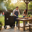 A man grills meat on a Myron Mixon Smokers Gravity Fed Smoker G20 in the backyard while a woman laughs and points at food nearby. Children play under glowing string lights, creating a cozy scene around the insulated smoker.