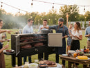 A group of adults enjoys barbecue on a grassy field, gathered around the Myron Mixon Gravity Fed Smoker RFG-60 by Myron Mixon Smokers. One man in an apron and hat grills meat while others eat, chat, and smile under string lights.