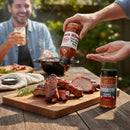 A person pours Myron Mixon Rib & Pork Kit barbecue sauce onto smoked ribs on a wooden board, with the kit’s rib rub seasoning nearby. In the background, someone smiles and enjoys a drink during an outdoor meal.