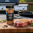 A jar of Myron Mixon Steak Seasoning by Myron Mixon rests on a wooden cutting board beside a raw steak topped with steakhouse flavor, fresh rosemary, and peppercorns, with a blurred grill and skillet in the background.