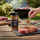 A hand sprinkles Myron Mixon Steak Seasoning onto a raw steak on a slate board, adding steakhouse-style flavor. A jar of Myron Mixon Steak Seasoning, utensils, another steak, and a fiery grill appear in the background.