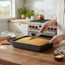 Someone slices a rectangular yellow cake in the DDR BBQ Supply 10 x 8-Inch Nonstick Baking Pan on a wooden kitchen counter, surrounded by baking ingredients and utensils for easy cleanup.