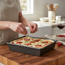 Someone sprinkles herbs on a rectangular pizza in the DDR BBQ Supply 10 x 8-Inch Nonstick Baking Pan atop a wooden countertop, with a glass of red wine and cherry tomatoes nearby, promising easy cleanup in a bright kitchen.