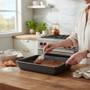 In a bright kitchen, someone spreads chocolate batter in a DDR BBQ Supply 10 x 8-Inch Nonstick Baking Pan. Eggs, flour, and a whisk rest on the wooden counter nearby for easy cleanup.