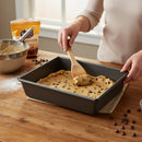 A person spreads cookie dough in a DDR BBQ Supply 10 x 8-Inch Nonstick Baking Pan with a wooden spoon. Nearby on the table are a bowl, whisk, and chocolate chips for easy cleanup.