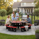 Five people gather outdoors on the Kanyon Living Poly Picnic Table Octagonal Red & Black, while one man grills nearby in a landscaped backyard with potted plants and a house in the background.