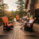 A man and a woman relax on a wooden deck furnished with Kanyon Living Adirondack Poly Glider Swivel Chairs in orange & black, beside a stone fireplace and potted plants, overlooking a forested landscape at sunset.
