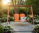 A Kanyon Living Poly Lumber 4′ Adirondack Swing in orange and black sits on a stone patio among greenery and flowers, topped with cushions and a blanket. String lights overhead add to the cozy outdoor setting at sunset.