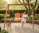 A woman in a light dress smiles and reads a book beside her golden retriever on the Kanyon Living Poly Lumber 4′ Adirondack Swing in orange and black, set in a sunlit garden with trees, flowers, and a small table holding a drink.