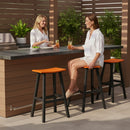Two women in white outfits enjoy drinks by an outdoor bar featuring a gray countertop, wooden panels, lush green plants, and Kanyon Living Poly Lumber Saddle Stool Bar Height Orange and Black by Kanyon Living.