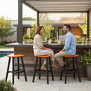 A man and a woman enjoy drinks and conversation on Kanyon Living Poly Lumber Saddle Stool Bar Height Orange and Black seating at an outdoor kitchen counter, with greenery, fruit, a house, and a pool in the background.