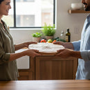 Two people stand in a kitchen, exchanging a round Words With Boards Seriously Kosher 14" personalized cutting board by Words with Boards, LLC. Fresh veggies and kitchen items in the background showcase thoughtful kosher kitchen organization.