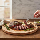 A hand lifts a slice of medium-rare steak with a fork from the Words With Boards Seriously Kosher Round Personalized Cutting Board (14" diameter) by Words with Boards, LLC; rosemary sprigs and green sauce sit nearby on a rustic wooden table.