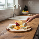 A hand slices blue cheese on the Words With Boards Seriously Kosher Round Personalized Cutting Board (14", Words with Boards, LLC), surrounded by assorted cheeses, crackers, and grapes on a wooden counter near a sink and fruit bowl.