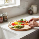 In a bright kitchen with jars by the window, someone slices carrots on the Words With Boards Seriously Kosher Round Personalized Cutting Board (14" diameter) along with assorted fresh veggies like tomatoes, cucumber, bell peppers, and broccoli.